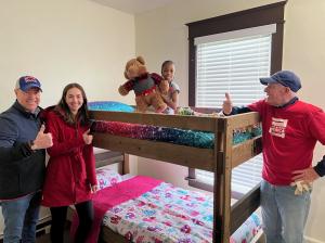 Three adults give thumbs up while standing next to a bunk bed with a child on the top bunk.