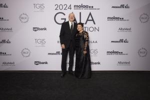 This image shows two individuals, a man and a woman, standing on a black carpet in front of a backdrop at an event. The backdrop displays the logos of various sponsors, including 